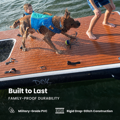 A dog wearing a blue life vest runs across a wooden deck on a floating platform in the water, with a child and another person partially visible. Text at the bottom reads 'Built to Last Family-Proof Durability' with feature callouts for military-grade PVC and rigid drop-stitch construction.